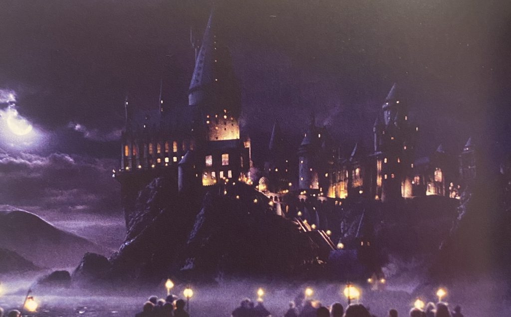 映画「ハリー・ポッターと賢者の石」のワンシーン。夜に浮かび上がる城、ホグワーツ魔法魔術学校。ボートに乗った生徒たちが向かっている。